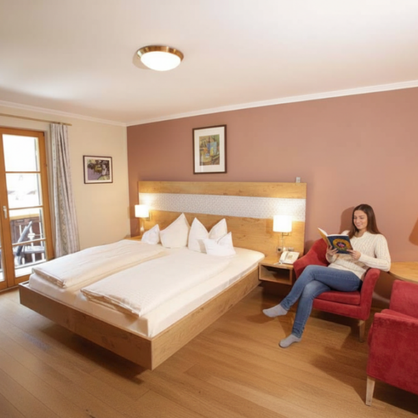 Gemütliches Zimmer im Klosterhof zur Post in Bayrischzell mit warmem Licht, Holzmöbeln und Sitzbereich. Eine Frau entspannt beim Lesen.