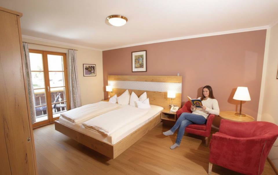 Gemütliches Zimmer im Klosterhof zur Post in Bayrischzell mit warmem Licht, Holzmöbeln und Sitzbereich. Eine Frau entspannt beim Lesen.