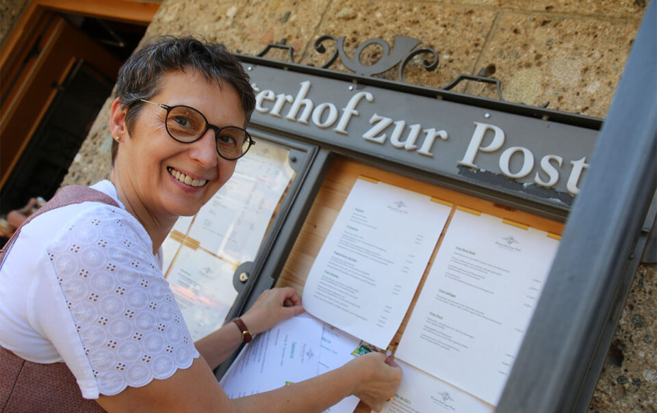 Viktoria Brummer befestigt lächelnd die neue Frühjahrs- und Sommer-Speisekarte im Schaukasten am Eingang des Klosterhofs zur Post in Bayrischzell.
