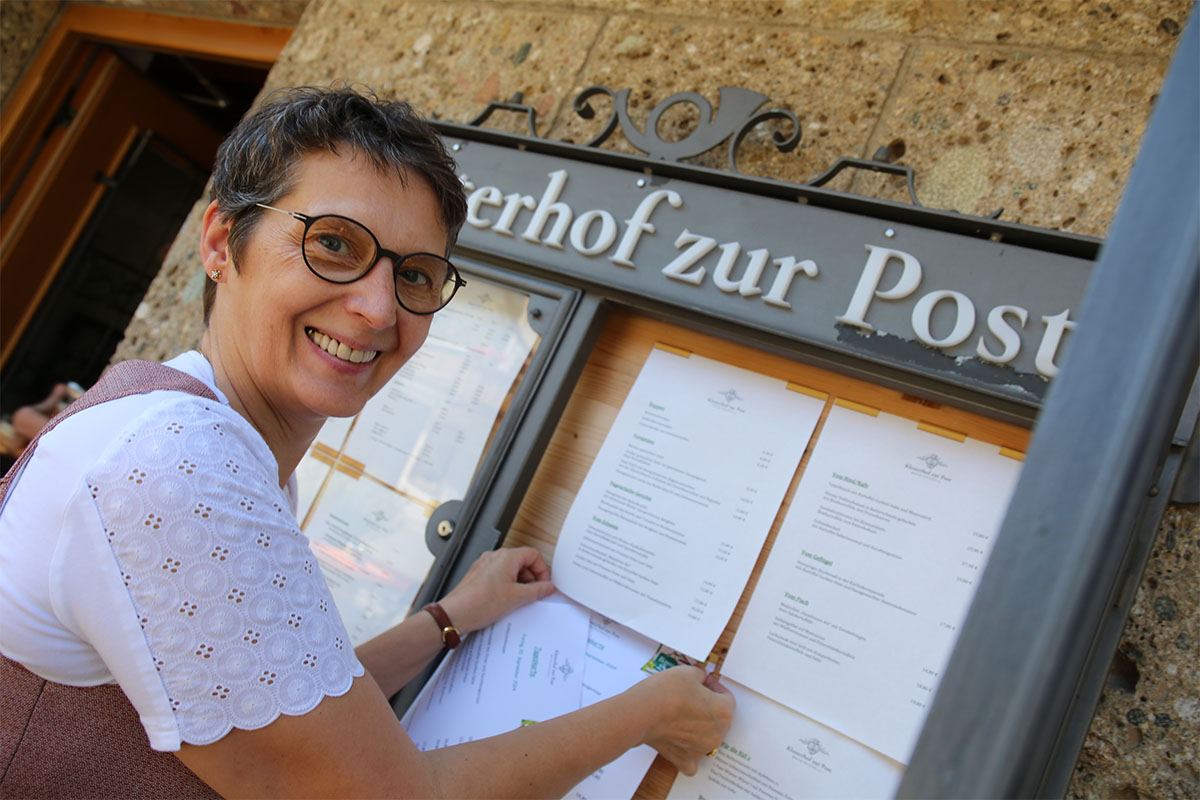 Viktoria Brummer befestigt lächelnd die neue Frühjahrs- und Sommer-Speisekarte im Schaukasten am Eingang des Klosterhofs zur Post in Bayrischzell.