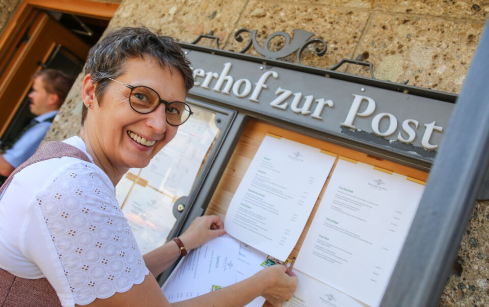 Viktoria Brummer befestigt lächelnd die neue Frühjahrs- und Sommer-Speisekarte im Schaukasten am Eingang des Klosterhofs zur Post in Bayrischzell.