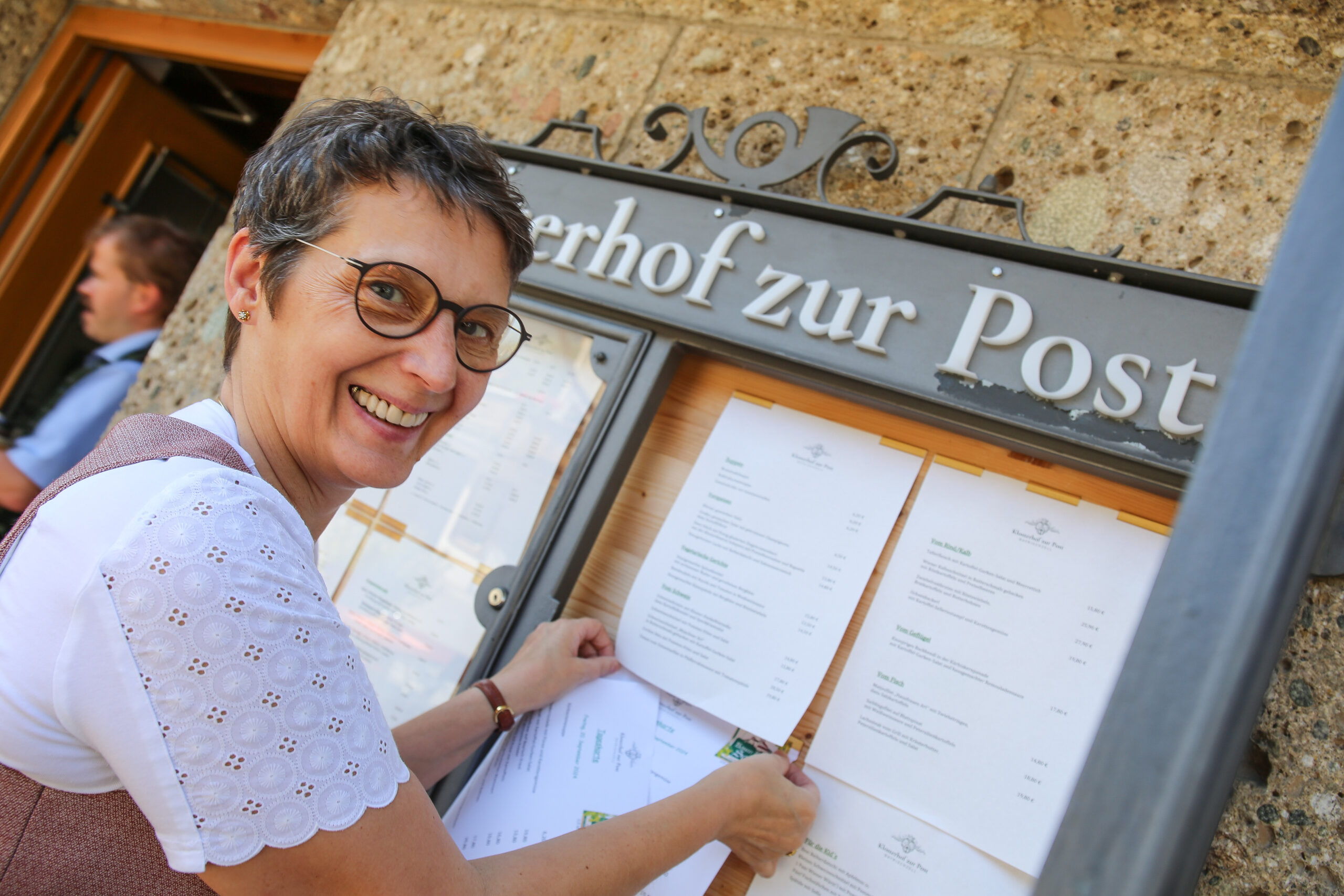 Viktoria Brummer befestigt lächelnd die neue Frühjahrs- und Sommer-Speisekarte im Schaukasten am Eingang des Klosterhofs zur Post in Bayrischzell.