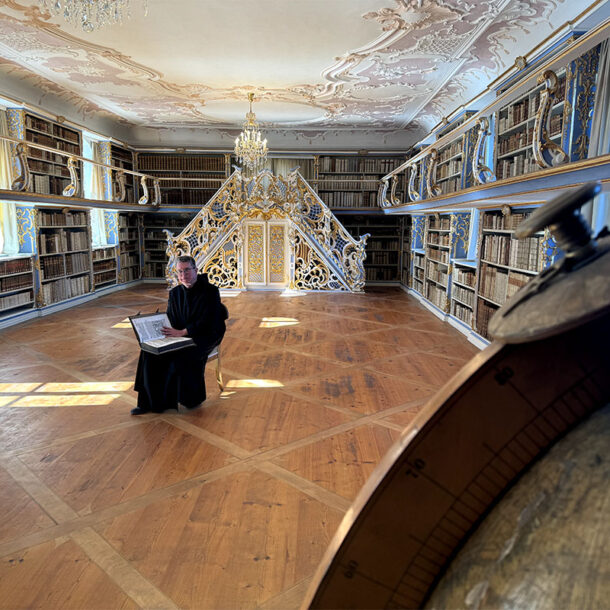 Historische Bibliothek des Klosters Scheyern mit Bücherregalen, Stuckdecke, Kronleuchtern und Pater Lukas mit einem Buch.