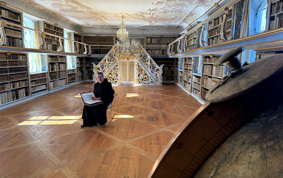 Historische Bibliothek des Klosters Scheyern mit Bücherregalen, Stuckdecke, Kronleuchtern und Pater Lukas mit einem Buch.