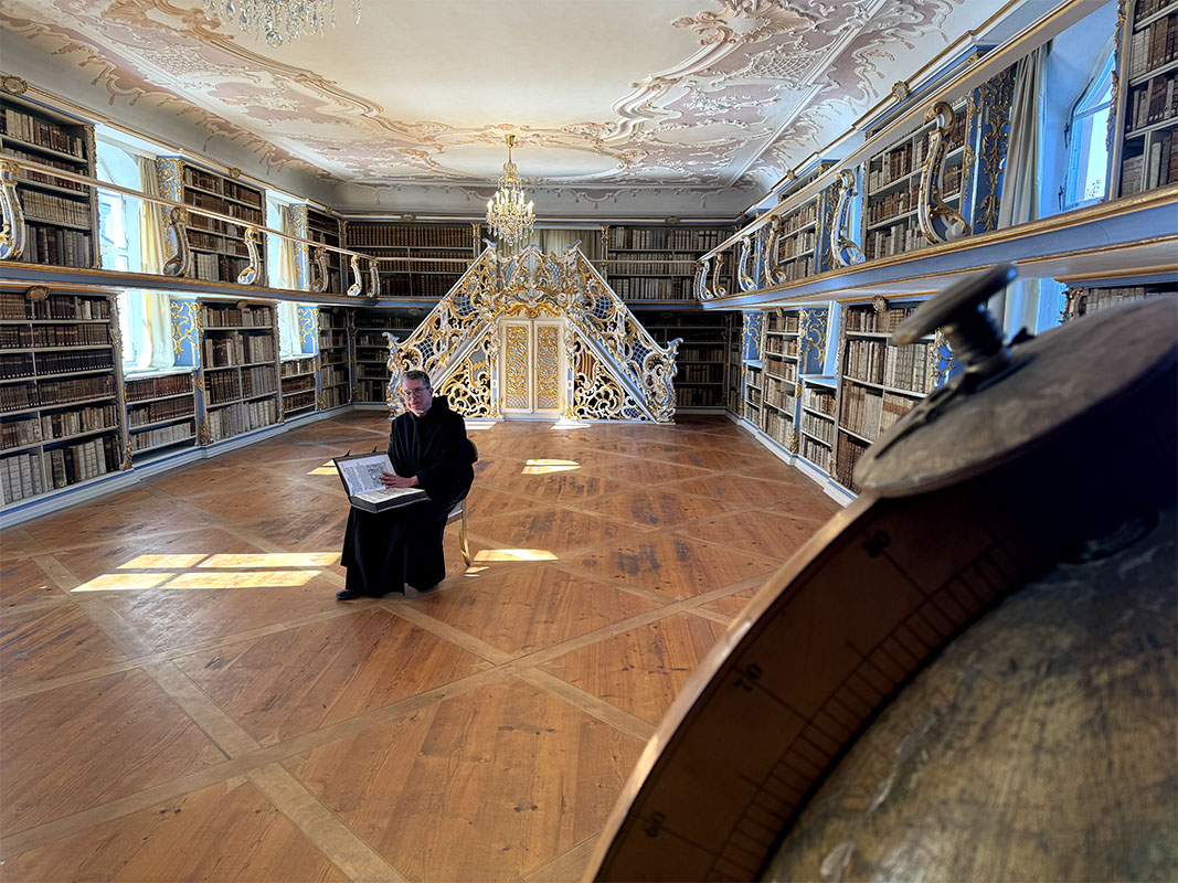 Historische Bibliothek des Klosters Scheyern mit Bücherregalen, Stuckdecke, Kronleuchtern und Pater Lukas mit einem Buch.