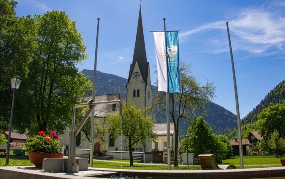 Pfarrkirche St. Margareth in Bayrischzell mit Jubiläumsfahne vor Bergkulisse und blauem Himmel – aufgenommen im Rahmen des Jubiläums 950 Jahre Bayrischzell und Kloster Scheyern.