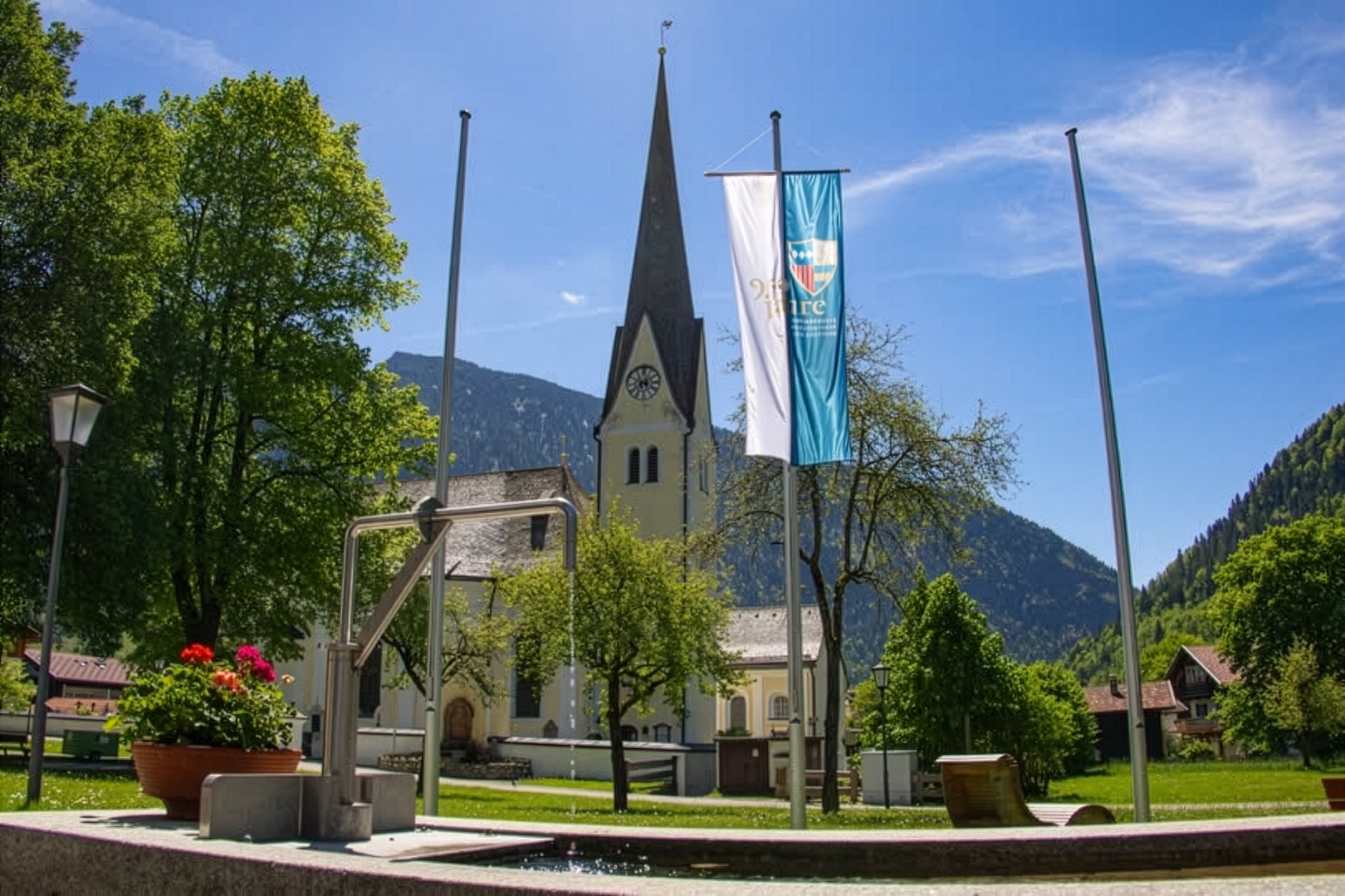 Pfarrkirche St. Margareth in Bayrischzell mit Jubiläumsfahne vor Bergkulisse und blauem Himmel – aufgenommen im Rahmen des Jubiläums 950 Jahre Bayrischzell und Kloster Scheyern.