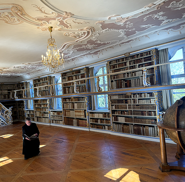 Pater Lukas sitzt in schwarzem Habit mit einem aufgeschlagenen Buch in der historischen Klosterbibliothek Scheyern. Umgeben von hohen Bücherregalen, barocker Stuckdecke, Kronleuchtern und einem großen Globus verbindet das Bild Wissen, Geschichte und klösterliche Tradition.