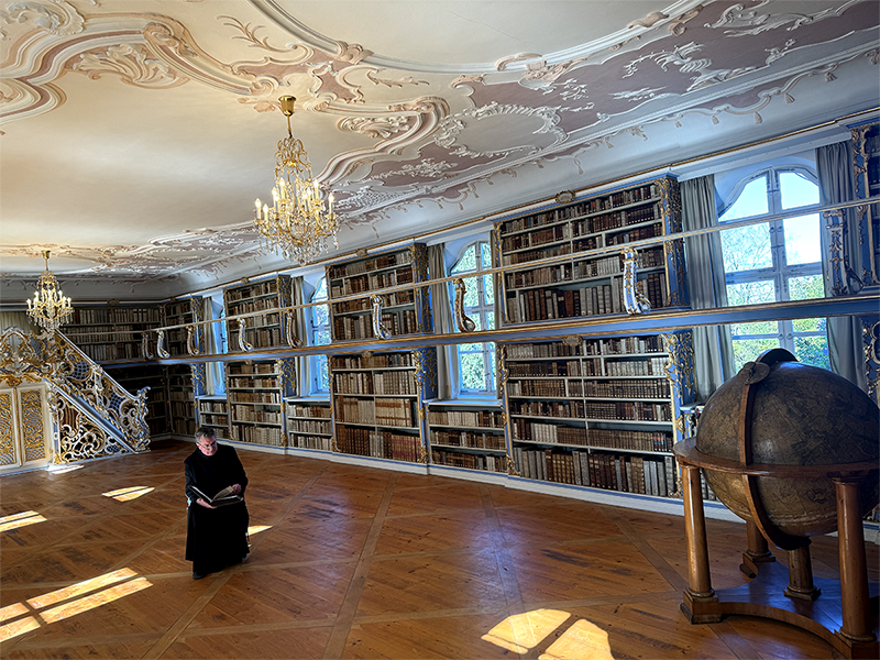 Pater Lukas sitzt in schwarzem Habit mit einem aufgeschlagenen Buch in der historischen Klosterbibliothek Scheyern. Umgeben von hohen Bücherregalen, barocker Stuckdecke, Kronleuchtern und einem großen Globus verbindet das Bild Wissen, Geschichte und klösterliche Tradition.